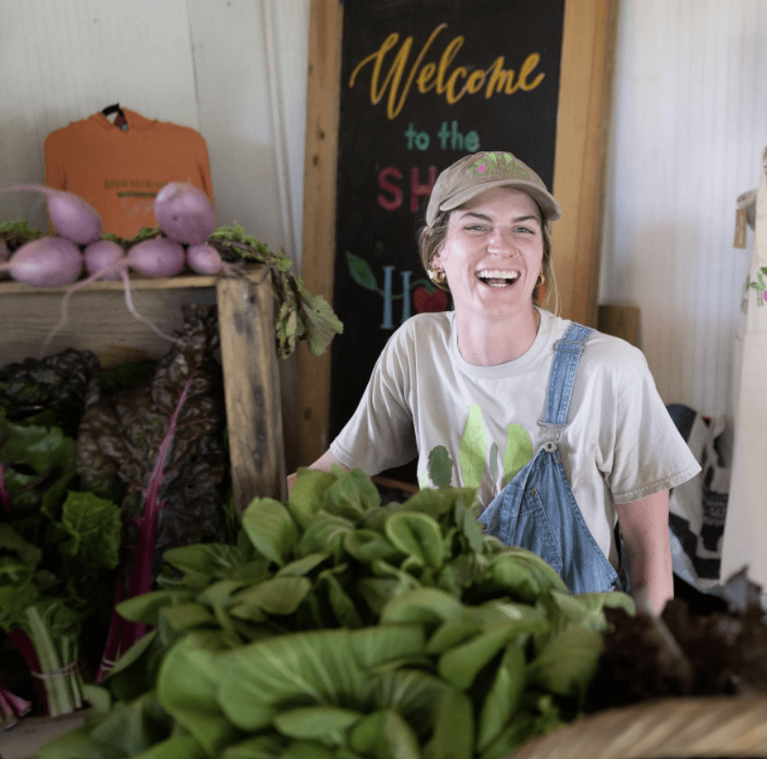 A woman smiling surrounded by a sign and produce such as lettuce.